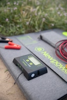 A digital solar charge controller with a display and buttons is placed on a gray mat with the brand name 'NewPower'. Red and black cables with clamps are also visible, set on a sandy surface with grassy vegetation in the background.
