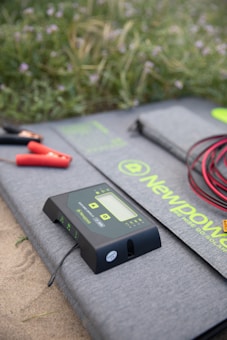 A digital solar charge controller with a display and buttons is placed on a gray mat with the brand name 'NewPower'. Red and black cables with clamps are also visible, set on a sandy surface with grassy vegetation in the background.