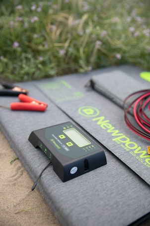 A digital solar charge controller with a display and buttons is placed on a gray mat with the brand name 'NewPower'. Red and black cables with clamps are also visible, set on a sandy surface with grassy vegetation in the background.
