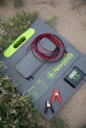 A solar panel lying on the ground in a sandy and grassy area. It has a grey fabric case with bright green accents and is marked with the brand name 'Newpowa 100W'. The panel is accompanied by a controller device, red and black cables, and two alligator clips. Wildflowers and grass are visible around the setup.