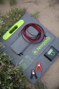 A solar panel lying on the ground in a sandy and grassy area. It has a grey fabric case with bright green accents and is marked with the brand name 'Newpowa 100W'. The panel is accompanied by a controller device, red and black cables, and two alligator clips. Wildflowers and grass are visible around the setup.