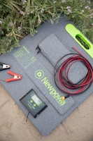 A solar panel kit is laid on the ground surrounded by green foliage with purple flowers. The kit includes a folded solar panel with a zippered pouch, a set of alligator clips, a charge controller, and coiled red and black cables. The brand 'Newpowa' is printed on the panel.