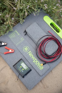 A solar panel kit is laid on the ground surrounded by green foliage with purple flowers. The kit includes a folded solar panel with a zippered pouch, a set of alligator clips, a charge controller, and coiled red and black cables. The brand 'Newpowa' is printed on the panel.