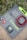 A solar panel kit is laid on the ground surrounded by green foliage with purple flowers. The kit includes a folded solar panel with a zippered pouch, a set of alligator clips, a charge controller, and coiled red and black cables. The brand 'Newpowa' is printed on the panel.