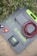 A solar panel kit is laid on the ground surrounded by green foliage with purple flowers. The kit includes a folded solar panel with a zippered pouch, a set of alligator clips, a charge controller, and coiled red and black cables. The brand 'Newpowa' is printed on the panel.