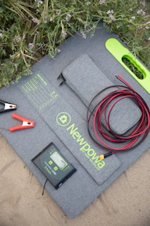 A solar panel kit is laid on the ground surrounded by green foliage with purple flowers. The kit includes a folded solar panel with a zippered pouch, a set of alligator clips, a charge controller, and coiled red and black cables. The brand 'Newpowa' is printed on the panel.