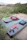 A grey case with the logo 'Newpowa' is lying outdoors on a sandy area surrounded by greenery. On the case are various objects including jumper cables with red and black clamps, a digital device with a screen, and a lime green zippered pouch.