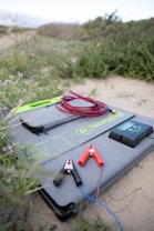 A grey case with the logo 'Newpowa' is lying outdoors on a sandy area surrounded by greenery. On the case are various objects including jumper cables with red and black clamps, a digital device with a screen, and a lime green zippered pouch.
