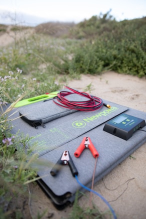 A grey case with the logo 'Newpowa' is lying outdoors on a sandy area surrounded by greenery. On the case are various objects including jumper cables with red and black clamps, a digital device with a screen, and a lime green zippered pouch.