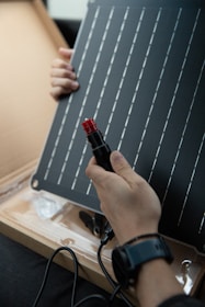 A person is holding a solar panel and an electrical connector. The panel features a grid of parallel lines, and the individual appears to be examining or assembling the components. The scene is indoors with the solar panel resting on a cardboard surface.