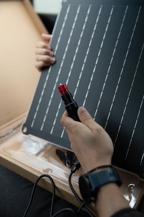 A person is holding a solar panel and an electrical connector. The panel features a grid of parallel lines, and the individual appears to be examining or assembling the components. The scene is indoors with the solar panel resting on a cardboard surface.
