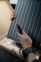 A person is holding a solar panel and an electrical connector. The panel features a grid of parallel lines, and the individual appears to be examining or assembling the components. The scene is indoors with the solar panel resting on a cardboard surface.