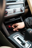 A hand is inserting or removing a red and black plug into a car's power socket located in the center console. The dashboard with various controls, including climate control knobs, is visible above.