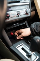 A hand is inserting or removing a red and black plug into a car's power socket located in the center console. The dashboard with various controls, including climate control knobs, is visible above.