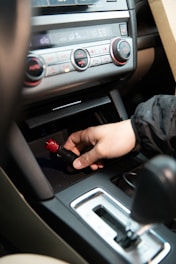 A hand is inserting or removing a red and black plug into a car's power socket located in the center console. The dashboard with various controls, including climate control knobs, is visible above.