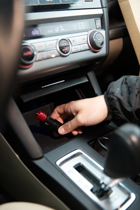 A hand is inserting or removing a red and black plug into a car's power socket located in the center console. The dashboard with various controls, including climate control knobs, is visible above.