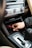 A hand is inserting or removing a red and black plug into a car's power socket located in the center console. The dashboard with various controls, including climate control knobs, is visible above.