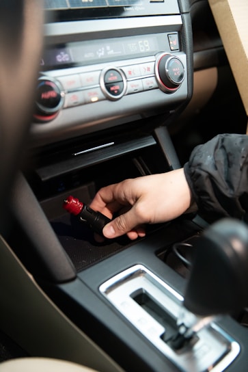 A hand is inserting or removing a red and black plug into a car's power socket located in the center console. The dashboard with various controls, including climate control knobs, is visible above.
