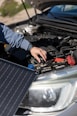 A person is adjusting or inspecting something under the hood of a car, with a focus on the battery and cables. A solar panel is positioned near the car, possibly for charging or testing purposes.