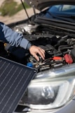 A person is adjusting or inspecting something under the hood of a car, with a focus on the battery and cables. A solar panel is positioned near the car, possibly for charging or testing purposes.