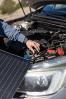 A person is adjusting or inspecting something under the hood of a car, with a focus on the battery and cables. A solar panel is positioned near the car, possibly for charging or testing purposes.