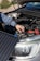 A person is adjusting or inspecting something under the hood of a car, with a focus on the battery and cables. A solar panel is positioned near the car, possibly for charging or testing purposes.