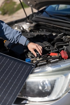 A person is adjusting or inspecting something under the hood of a car, with a focus on the battery and cables. A solar panel is positioned near the car, possibly for charging or testing purposes.