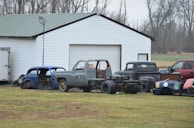 A collection of vintage car and truck bodies is scattered in front of a large white garage with a green metal roof. The scene is set on a grassy field with leafless trees in the background, indicating a late autumn or winter season. Various car parts, including an old blue car body and a gray truck, are visible. The vehicles appear to be in various stages of disrepair or restoration.