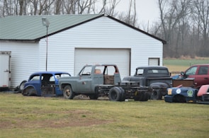 A collection of vintage car and truck bodies is scattered in front of a large white garage with a green metal roof. The scene is set on a grassy field with leafless trees in the background, indicating a late autumn or winter season. Various car parts, including an old blue car body and a gray truck, are visible. The vehicles appear to be in various stages of disrepair or restoration.
