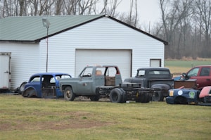A collection of vintage car and truck bodies is scattered in front of a large white garage with a green metal roof. The scene is set on a grassy field with leafless trees in the background, indicating a late autumn or winter season. Various car parts, including an old blue car body and a gray truck, are visible. The vehicles appear to be in various stages of disrepair or restoration.