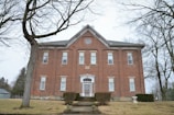 A large, two-story red brick building with white-framed windows and a peaked roof. There is a symmetrical arrangement of windows, with a prominent central doorway flanked by small bushes. The building is surrounded by bare trees and a patchy lawn.