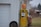 An old, yellow Shell gas pump is positioned next to a white metal building with a closed garage door. A spare tire is leaning against the wall near the pump. The setting appears rustic, with overgrown grass and an old, weathered aesthetic.