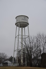 A tall, white water tower with the text 'ST. PARIS' written on the tank. The structure is supported by metal beams and set against a gray, overcast sky. Bare trees and residential houses are visible at the base of the tower.