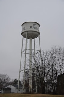 A tall, white water tower with the text 'ST. PARIS' written on the tank. The structure is supported by metal beams and set against a gray, overcast sky. Bare trees and residential houses are visible at the base of the tower.