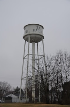 A tall, white water tower with the text 'ST. PARIS' written on the tank. The structure is supported by metal beams and set against a gray, overcast sky. Bare trees and residential houses are visible at the base of the tower.