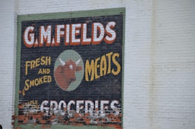 A vintage-style advertisement painted on a brick wall features large text that reads 'G.M. Fields Fresh and Smoked Meats' accompanied by an illustration of a cow's head. The sign also mentions 'Groceries' at the bottom.