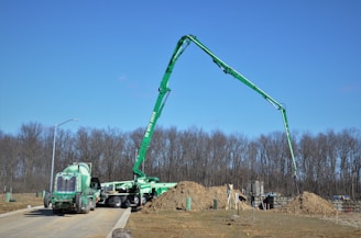 A green construction truck with an extended boom is situated at a construction site with piles of dirt and unfinished structures. Leafless trees are in the background under a clear blue sky.