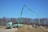 A green construction truck with an extended boom is situated at a construction site with piles of dirt and unfinished structures. Leafless trees are in the background under a clear blue sky.