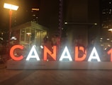 Large illuminated letters spelling 'CANADA' are displayed in an urban setting at night. Several people sit or stand on the letters, with city lights visible in the background.