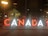 Large illuminated letters spelling 'CANADA' are displayed in an urban setting at night. Several people sit or stand on the letters, with city lights visible in the background.
