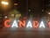 Large illuminated letters spelling 'CANADA' are displayed in an urban setting at night. Several people sit or stand on the letters, with city lights visible in the background.