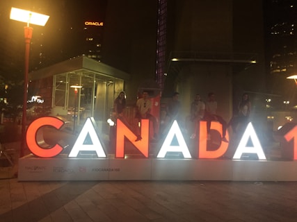 Large illuminated letters spelling 'CANADA' are displayed in an urban setting at night. Several people sit or stand on the letters, with city lights visible in the background.