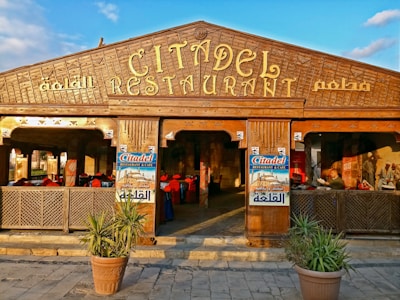 A wooden facade of a restaurant with intricate carvings and large signs displaying 'Citadel Restaurant & Cafe'. Potted plants flank the entrance, and the decor inside includes red chairs and murals on the walls. A person can be seen reading.