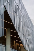 A modern architectural facade is characterized by vertical metal slats that create a textured, geometric pattern. Behind these slats, the building's interior is visible with warm lighting, revealing a concrete support column and glass elements. The design features clean lines and an industrial aesthetic.