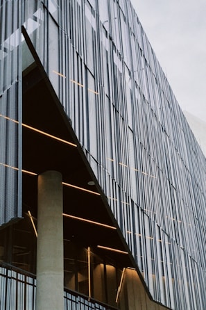 A modern architectural facade is characterized by vertical metal slats that create a textured, geometric pattern. Behind these slats, the building's interior is visible with warm lighting, revealing a concrete support column and glass elements. The design features clean lines and an industrial aesthetic.
