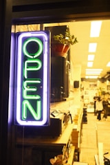 A neon sign with the word 'OPEN' in bright green is prominently displayed on the left. It is illuminated against a backdrop of a cozy, warmly lit interior, which appears to be a shop or cafe. Inside, shelves are filled with various items, and a plant is visible in a pot on a shelf. There are a few people visible in the background, contributing to a lively atmosphere.