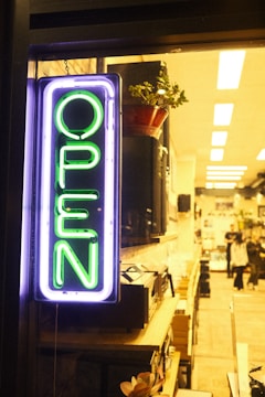 A neon sign with the word 'OPEN' in bright green is prominently displayed on the left. It is illuminated against a backdrop of a cozy, warmly lit interior, which appears to be a shop or cafe. Inside, shelves are filled with various items, and a plant is visible in a pot on a shelf. There are a few people visible in the background, contributing to a lively atmosphere.