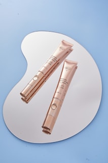 Two elegant gold-colored tubes of eye cream placed on a reflective, irregularly shaped surface. The background is a solid light blue color, enhancing the sophisticated appearance of the products.