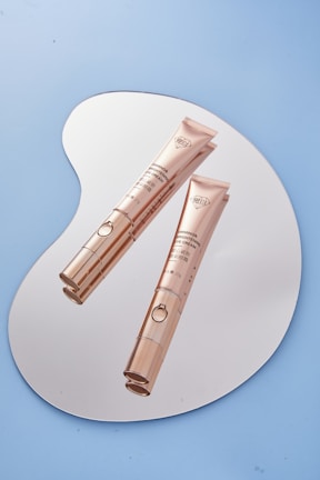 Two elegant gold-colored tubes of eye cream placed on a reflective, irregularly shaped surface. The background is a solid light blue color, enhancing the sophisticated appearance of the products.