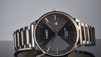A sophisticated wristwatch with a black dial and rose gold markers, featuring the brand name 'Timex' and 'Sapphire Crystal' on the face. The stainless steel strap complements the minimalist and elegant design, with a small date window displaying the number two.